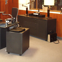 Wnętrze biura w kolorach brązu. Na podłodze jasno-beżowa wykładzina. Fotel przy biurku ze skórzaną, ciemno-brązową tapicerką.